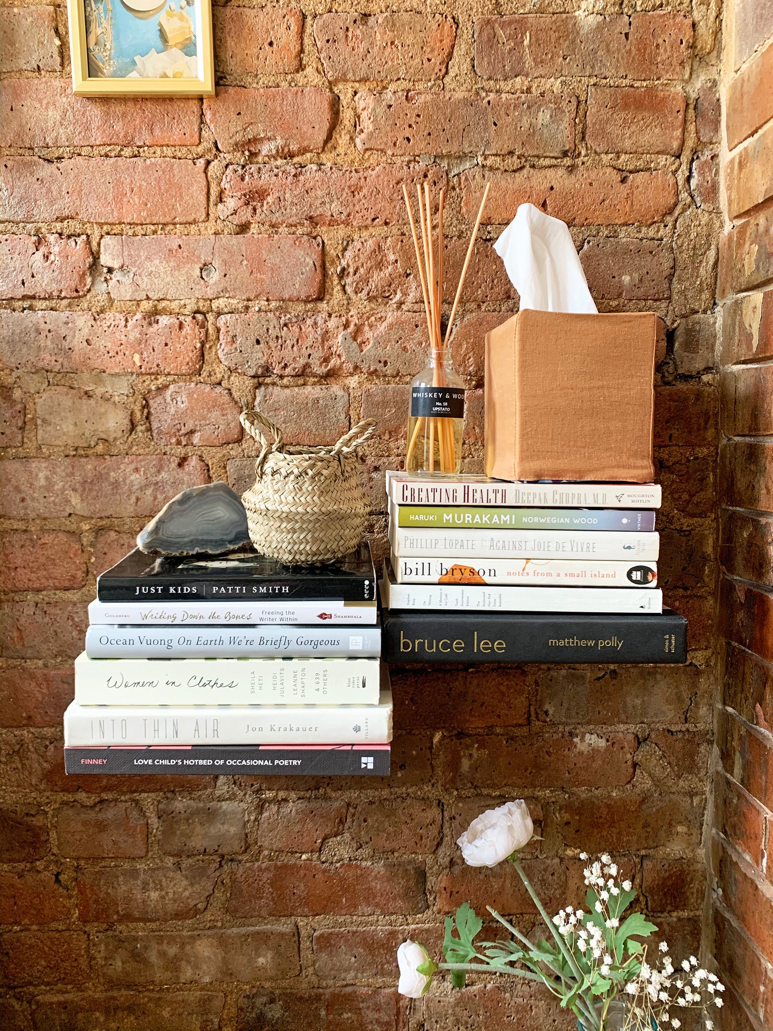 A brick wall with two shelves made out of stacks of books with a stone and a bamboo basket on top on one stack and an oil diffuser and a tissue box with an orange linen tissue box cover on top of the other.