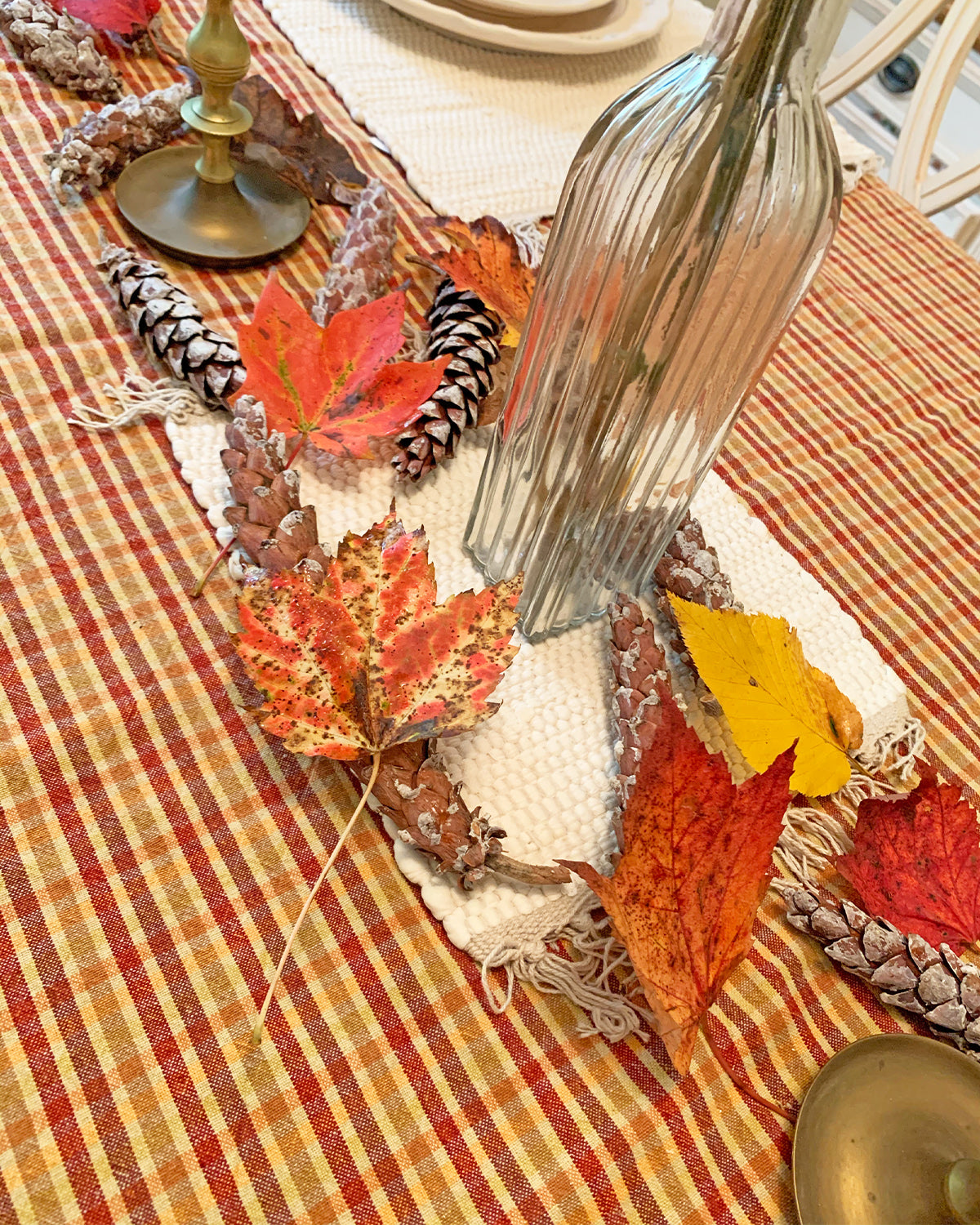 Closeup of table scape with orange and red checkered tablecloth, small white rug hot pad under neath a cluster of red and orange leaves and pinecones, a glass bottle in center filled with dried flowers.