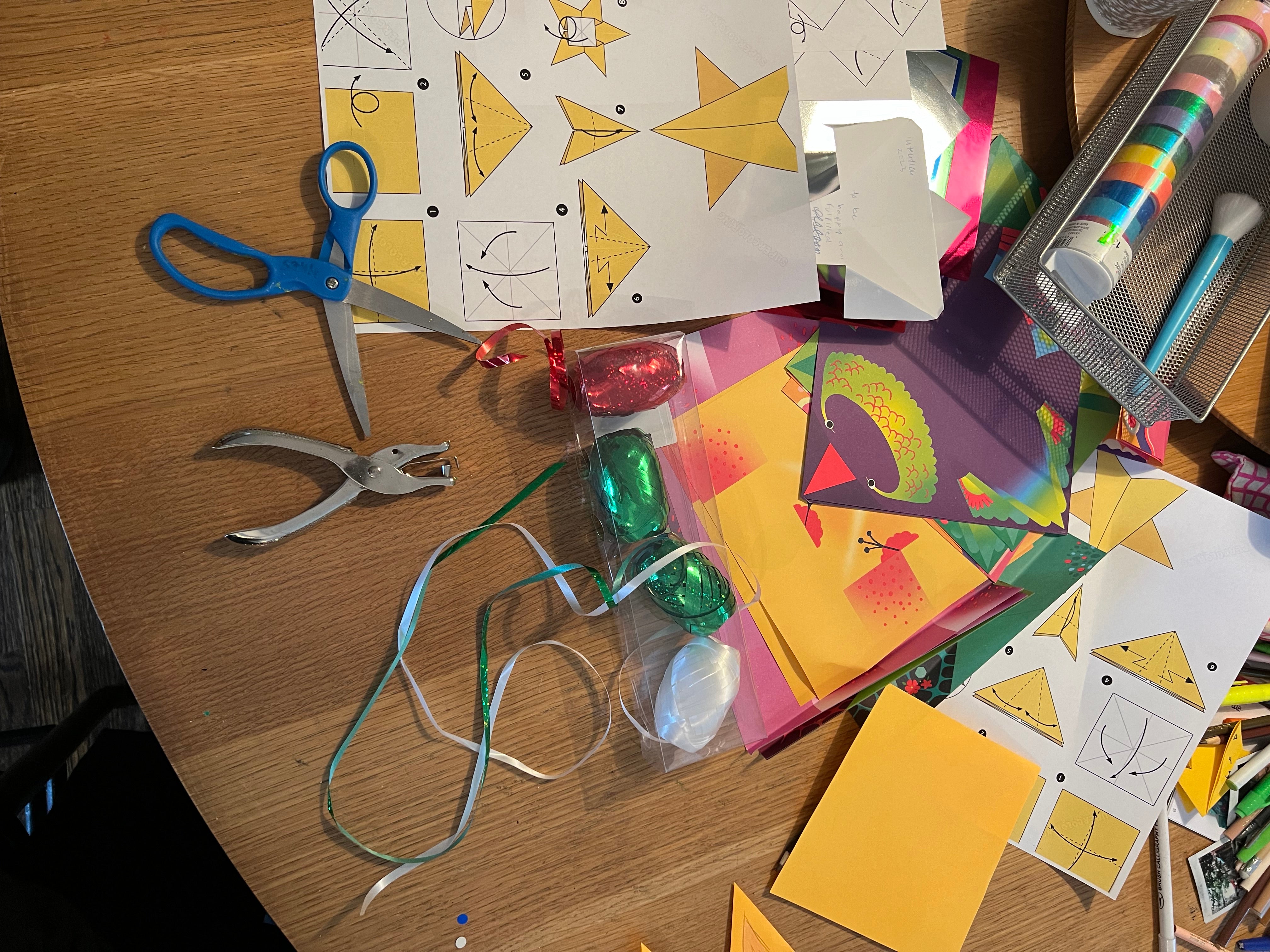 Image of scissors, hand held hole punch, Christmas gift wrap ribbon, printed origami instructions and other various craft supplies laying on a table