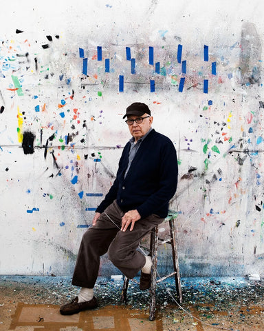 American artist, ellsworth kelly, sitting on a stool posing in front of one of his paintings