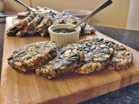 Vegan raw seed bread on a wooden cutting board with a ramekin filled with pesto and a teaspoon in the background.