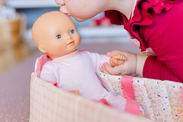 Toddler kissing pomea doll on head as it sits in a bassinet