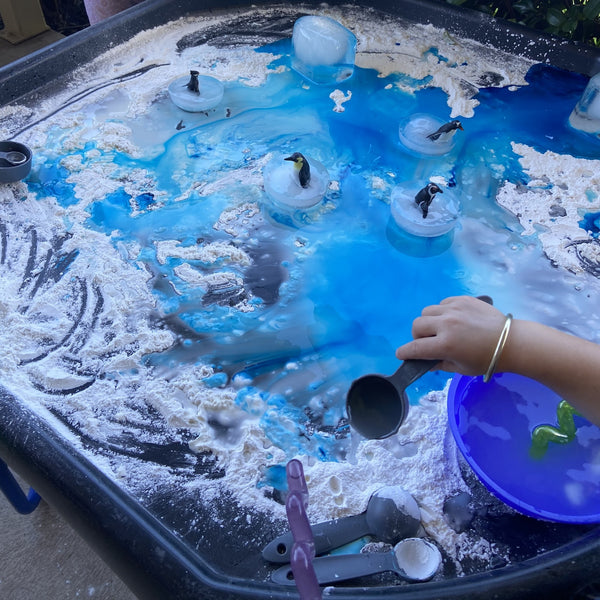 Tuff tray with frozen penguins and corn flour on it with food colouring drops and kids playing and making messy water play