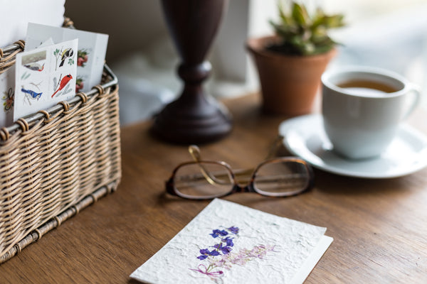 a desk top holding a basket with stamps, a card, a pair of reading glasses, a lamp and a cup of coffee