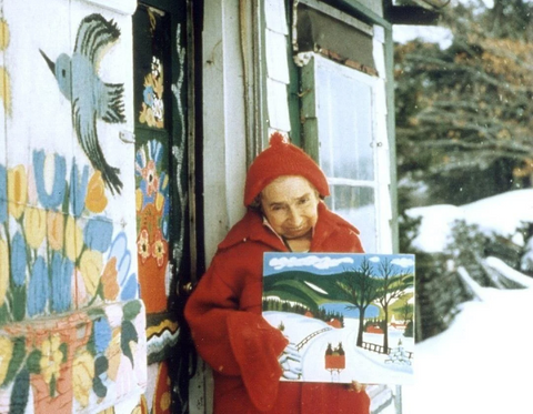Maud Lewis - holding one of her art pieces out in front of her house.