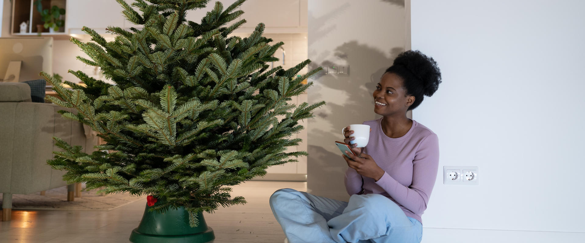 A woman with a mug sat on the floor looking up at an undecorated Christmas tree