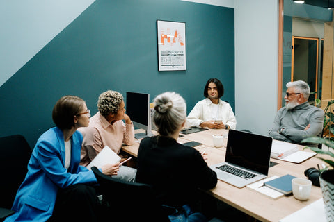 Group of diverse professionals seated around a table in a modern office space, engaged in discussion. A poster promoting 'Handmade-Arcade' is visible on the wall. They are surrounded by office essentials like laptops, mugs, and notepads.