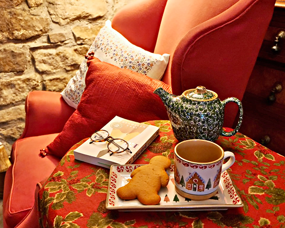 Cosy Reading nook with armchair Nicholas Mosse Linen cushions, Nicholas Mosse Pottery teapot, large mug and rectangular plate with gingerbread man biscuit. Book with glasses on table beside.