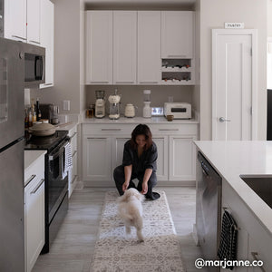 Emile latte neutral floral print standing mat shown in white kitchen with woman bending down on the mat and little white dog running toward her