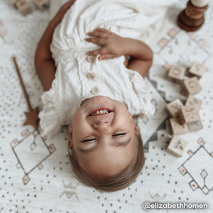 Neutral boho print baby play mat shown with baby girl laying on floor with toys. @elizabethhomen written in bottom right hand corner.