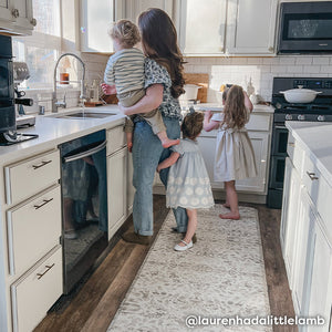 Emile latte neutral floral print standing mat shown in size 30x108 with Mom standing on the mat in the kitchen holding her toddler son and 2 daughters surrounding her