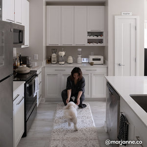 Emile latte neutral floral print standing mat shown in white kitchen with woman bending down on the mat and little white dog running toward her