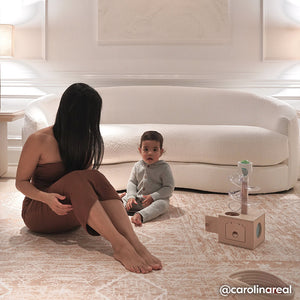 Ula amber brown boho print play mat shown in living room with white couch and mom and baby playing with jack in the box toy on the mat. @carolinareal written in bottom right hand corner.