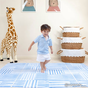 hadley french blue inverted stripe blue and white tumbling mat shown in a plat room with stacked baskets and giant giraffe plush toy with toddler boy running across the mat. @anaperu written in the lower left hand corner