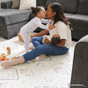 Neutral boho print baby play mat shown in play room with a mom holding a baby and kissing her toddler. @adrileon_ written in bottom right hand corner.