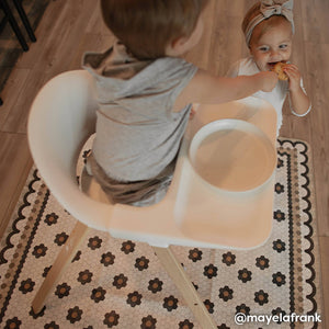 Daisy charcoal penny tile print highchair mat shown with baby boy in highchair and little sister eating a cookie standing below him