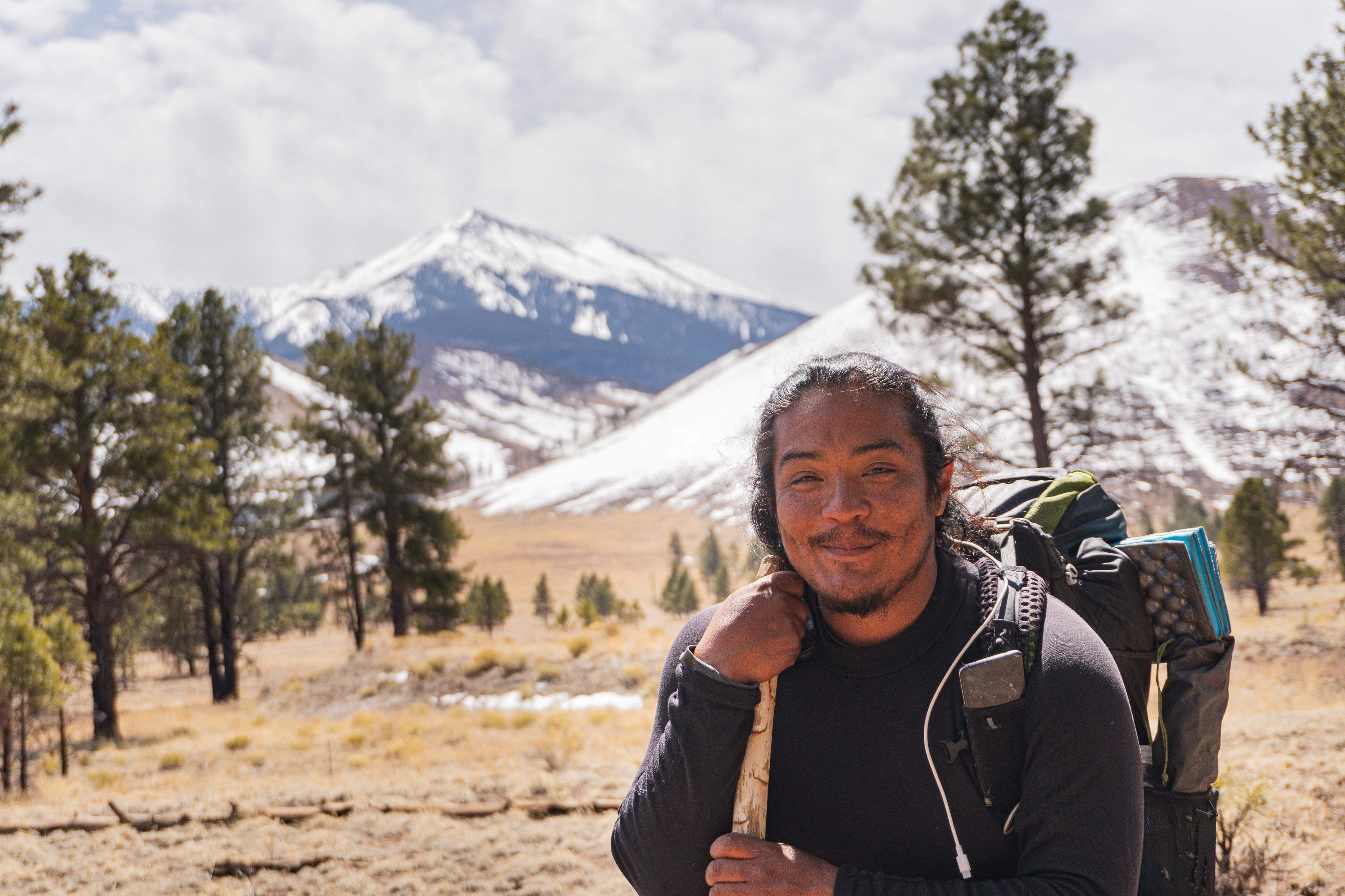 Gabe Vasquez smiling at the camera with a backpacking backpack and a walking stick in hand.