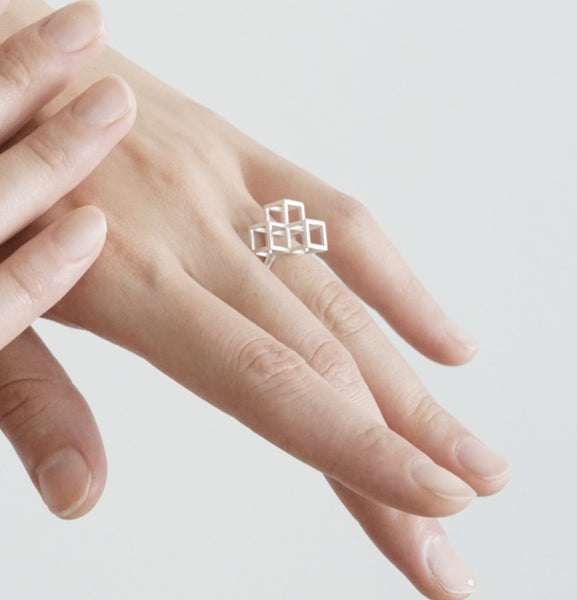 A silver ring with three cubes sits on the finger of a woman's hand in front of a light grey background