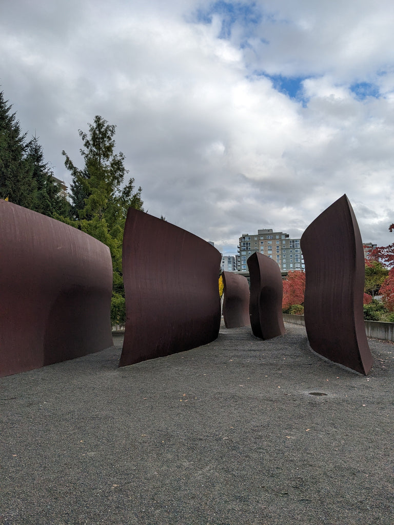 Four large panels of brown steel stand on the grey ground in front of trees and a cloudy sky.