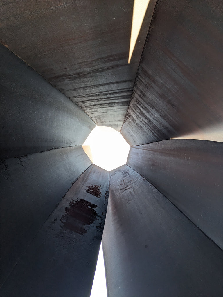 A heptagonal hole opens up to the white sky at the top of a large brown steel sculpture.