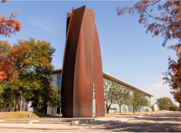 A tall brown steel sculpture stands in front of a building surrounded by trees and with a blue sky in the background