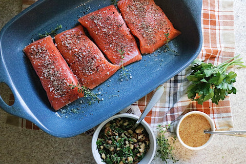 Raw salmon seasoned in baking dish next to small bowls of toppings