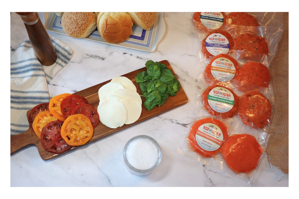 Fresh tomatoes, mozzarella, and basil on a wooden board and surrounded by all of our salmon burgers, buns, salt and pepper