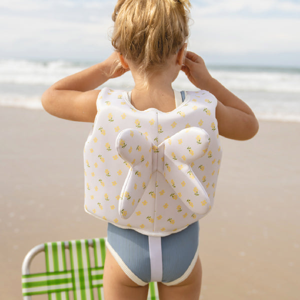 Image shows a child in a blue swim suit wearing a Mima Swim Vest looking out to the sea., we can see the back of her swim vest