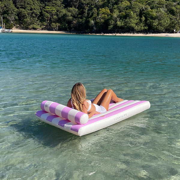Blonde woman reclines on the Luxe Lilo Pink Stripe floating on a lake