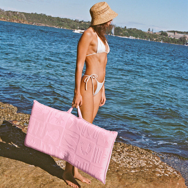 Young woman in a white bikini stands beside the water holding the Terry Folding Seat De Playa Pink by the carry handle