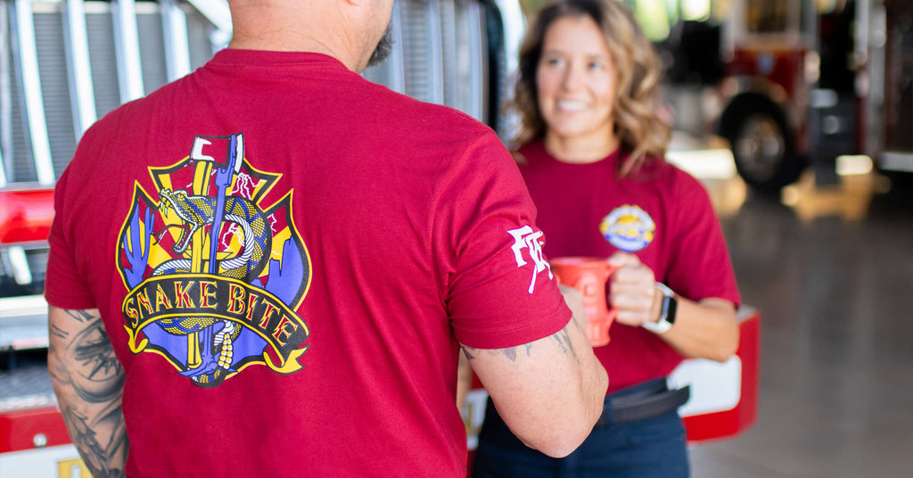 Two firefighters standing together, talking at a fire station while drinking coffee.