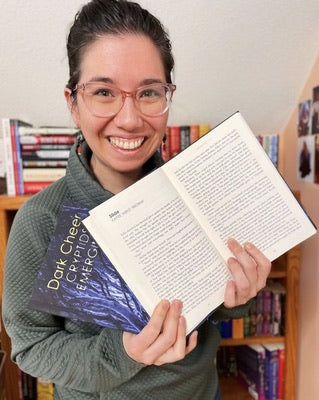 [IMAGE ID: Kaitee (light skinned, mixed race Asian woman with dark hair, pulled up, and glasses) smiles at the camera, an open book in her hand. It is open to a page titled “Slide” with Kaitee Yaeko Tredway under it.]