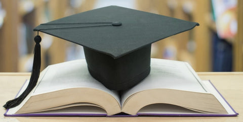 The shelves of a library in the background; in the foreground a very thick book open in thee middle with a graduates black cap and tassel resting on it