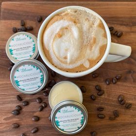 Three tins of Mochalicious body butter with a cup of frothy coffee and coffee beans scattered on a table