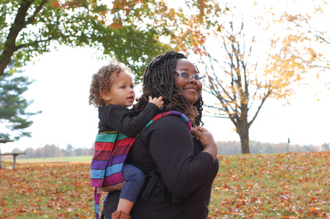 Woman with baby in a multicolor striped baby carrier. Baby is worn on woman's back.