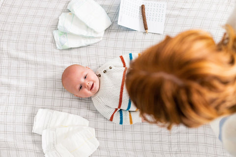 Baby laying on floor with diapers surrounding his head with red-haired woman leaning over him 