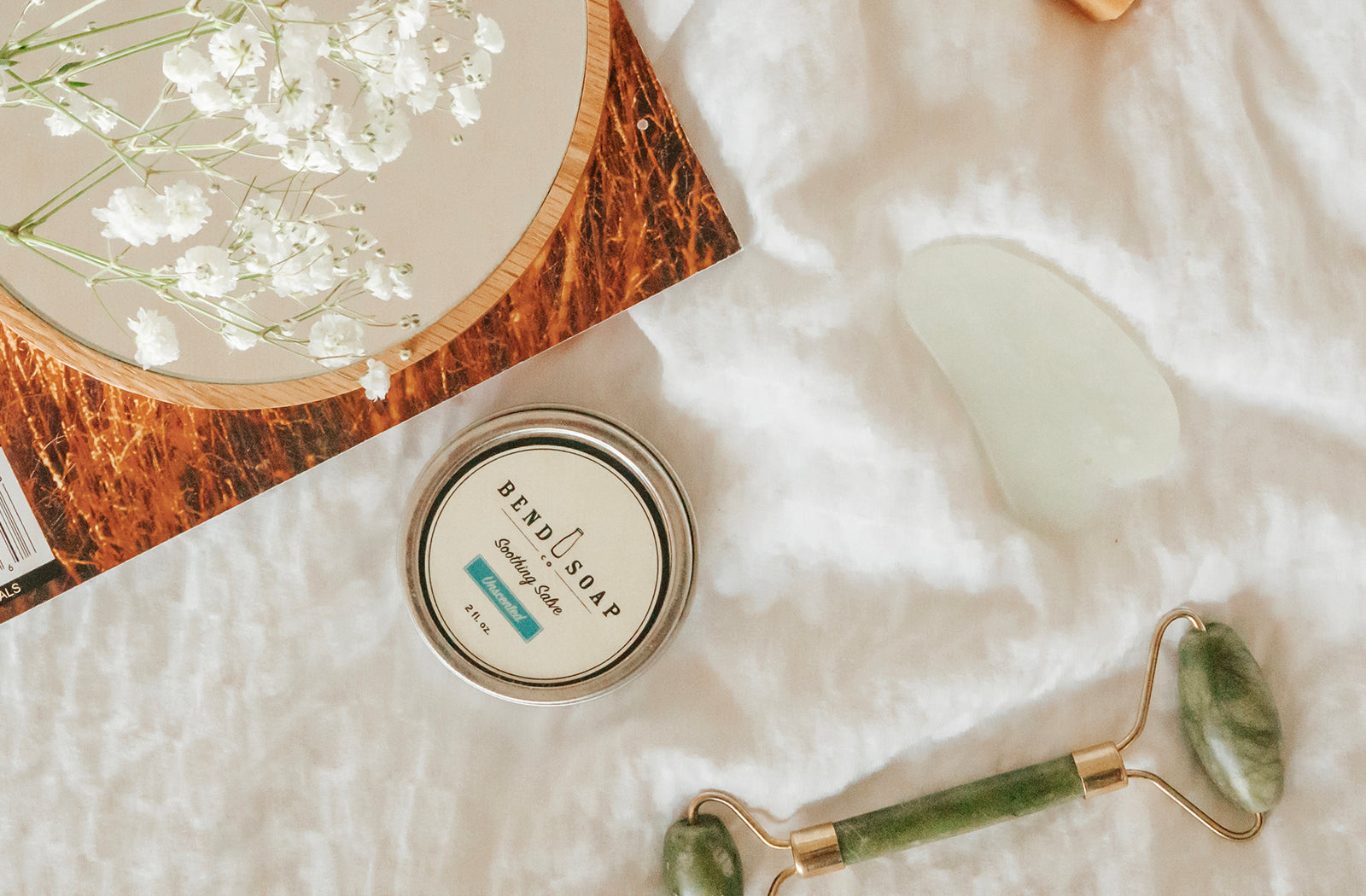 a flat lay of flowers, face roller, and all natural salve on a white comforter