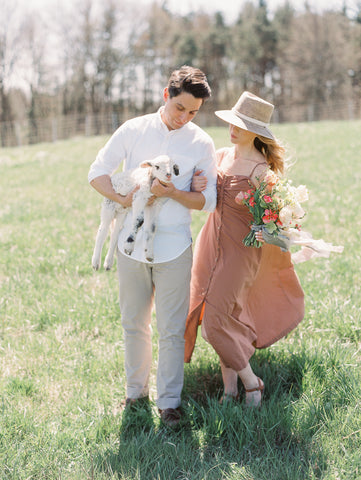 Engagement photo of couple in a field holding a lamb and a spring bouquet with silk ribbon by The Lesser Bear Photo by Jenny Haas Florals Old Slate Farm Design and Planning Auburn and Ivory Creative