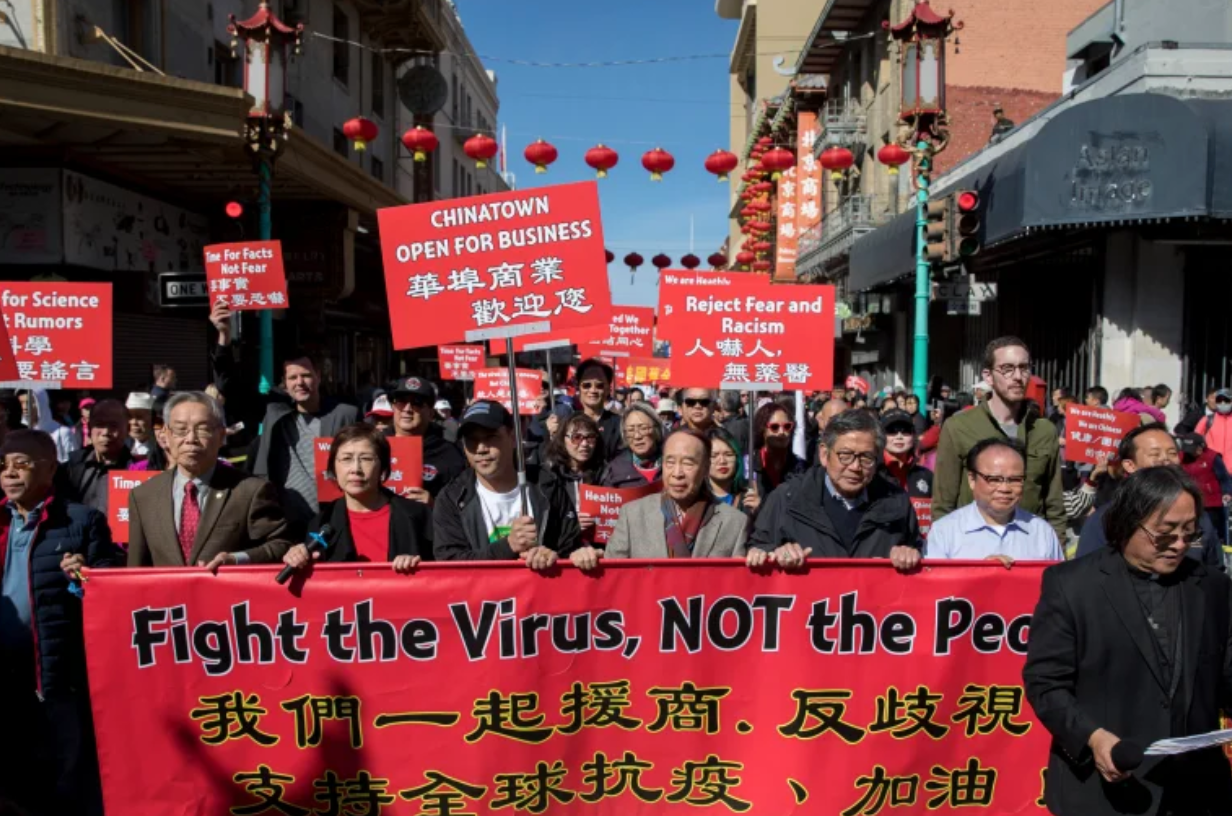 Photo of crowd in Chinatown holding a sign that says "Fight the virus, NOT the people."