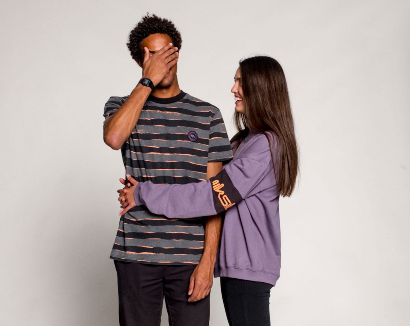 Man covering his face wearing a grey tshirt with black stripes hugged by girl wearing a purple sweater 