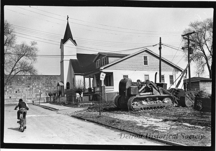 Black and white photographic print mounted on board depicting Holy Trinity Lutheran Church at 6570 Elmwood. The Cadillac Assembly Plant can be seen in the left of the background. A house can be seen to the right of the church, surrounded by a fence. A bulldozer can be seen in the foreground and a child is riding a bike down the street. Dated April 1981.