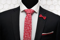 An outfit image featuring a black blazer and white button down shirt. The lapel pin is a bow shaped red polka dot pin and is paired with a red silk knitted pocket square, and the red Shakespeare floral skinny red tie.