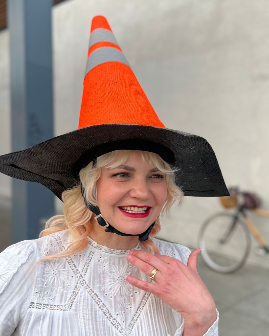 A joyful blonde woman, donning a whimsical helmet resembling a traffic cone, flashes a radiant smile. In the softly blurred background, her bicycle leans against a weathered white wall, adding a sense of adventure to the scene.