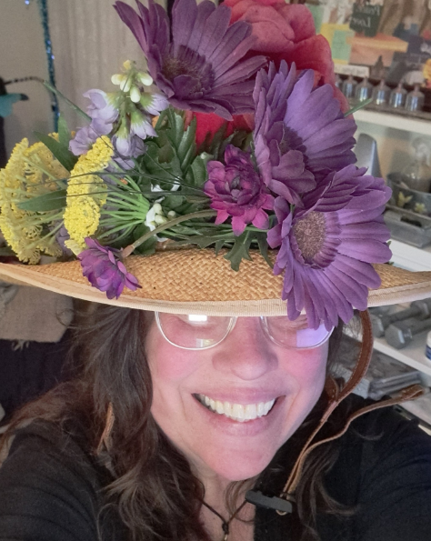 A smiling woman wears a decorated straw hat covered in purple, red, and yellow blossoms.