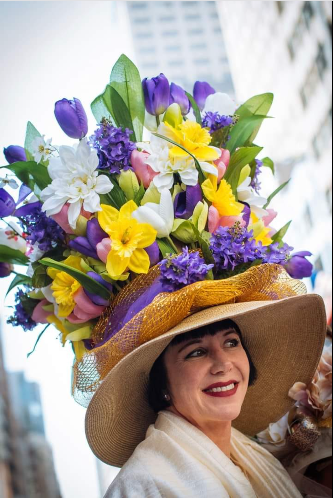 An absolute unit of a decorated easter bonnet. The crown of wide brimmed straw hat is completely obscured by a tower of purple, white, and yellow flowers.