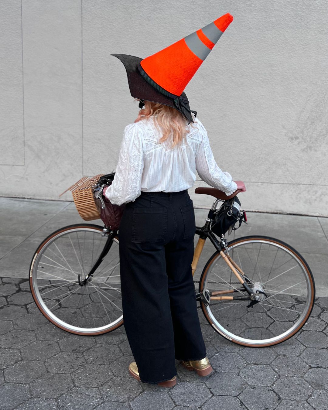 A woman with blonde hair, captured from behind as she strolls beside a weathered white wall, guides her bicycle by her side. She dons an eye-catching bicycle helmet, uniquely covered to mimic the appearance of a fluorescent orange traffic cone, adding a whimsical yet safety-conscious vibe to the scene.