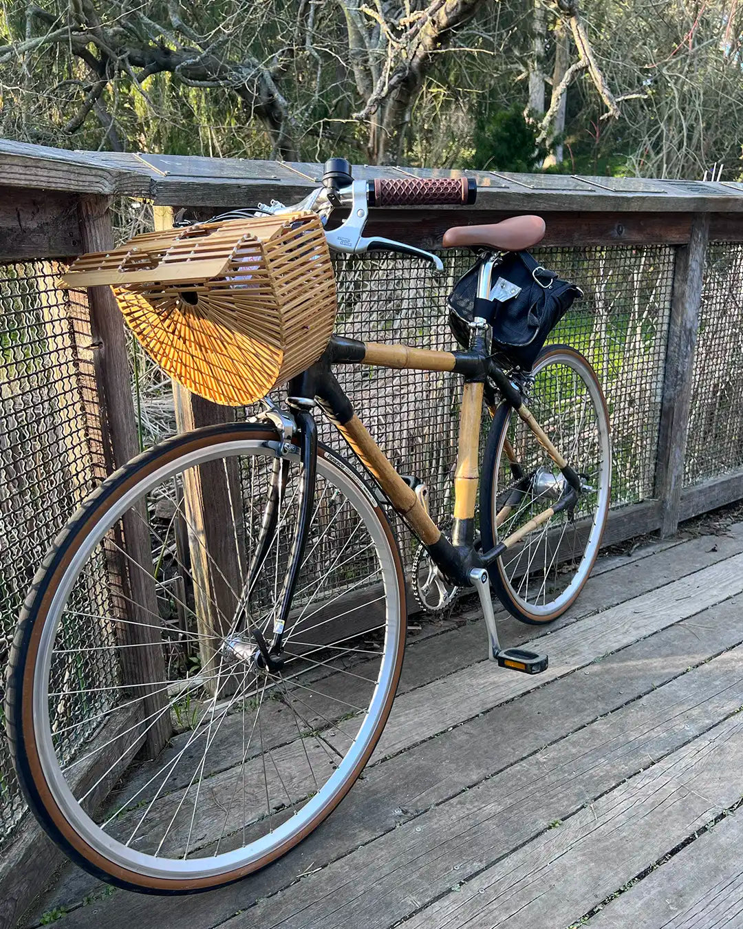 A bamboo bicycle with a stylish bamboo bike basket mounted on the front, parked on a wooden bridge in natural light.