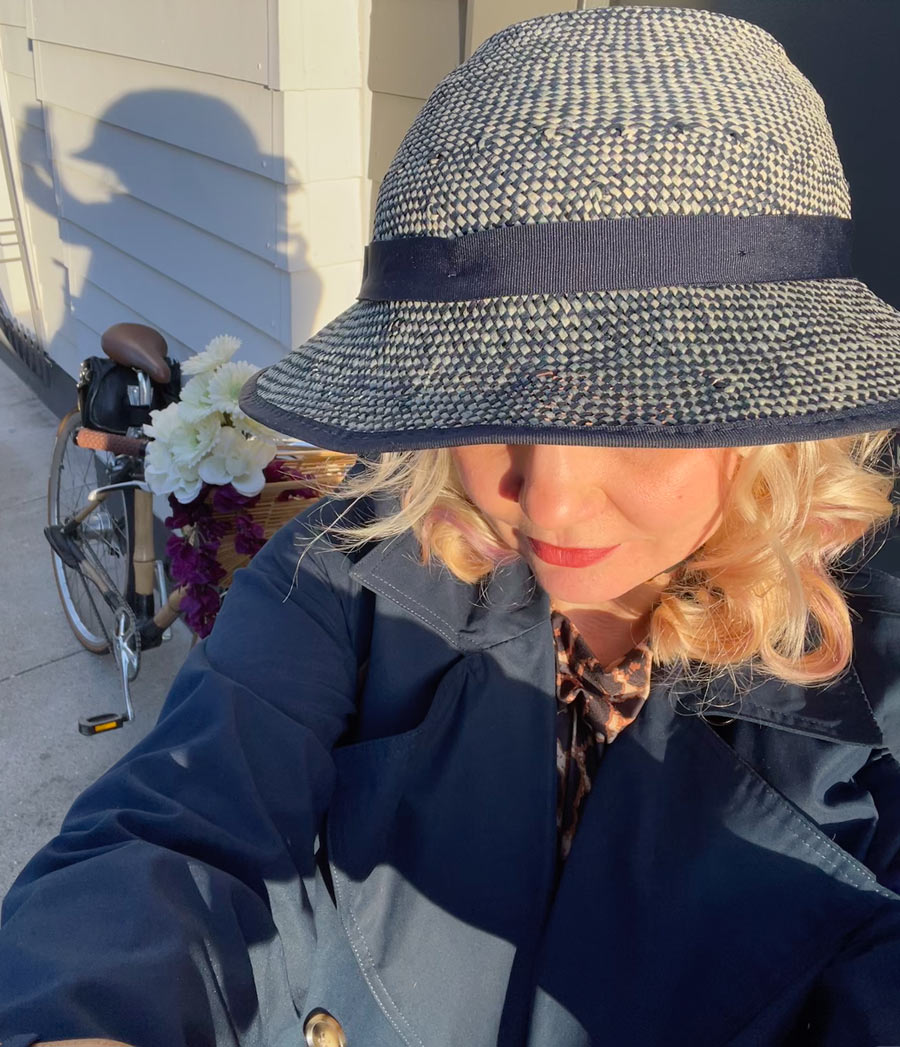 Woman in a Shibori Blue Straw Hat Bike Helmet and a navy blue trench coat poses in the sun with a bicycle in the background.