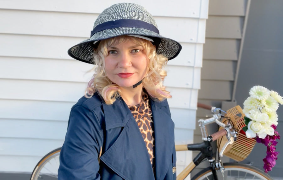 Melissa wears the Thousand Helmet with the Shibori Blue Straw Hat Helmet cover on top. A bicycle is visible in the background.