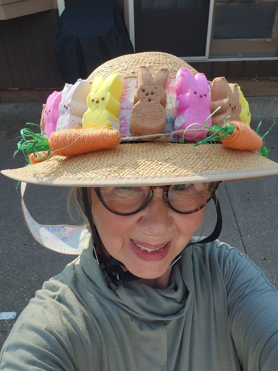 A smiling lady radiates good vibes from underneath her decorated Straw Hat Bike Helmet. Her bonnet is festooned with a garland of peeps and carrots made of yarn.